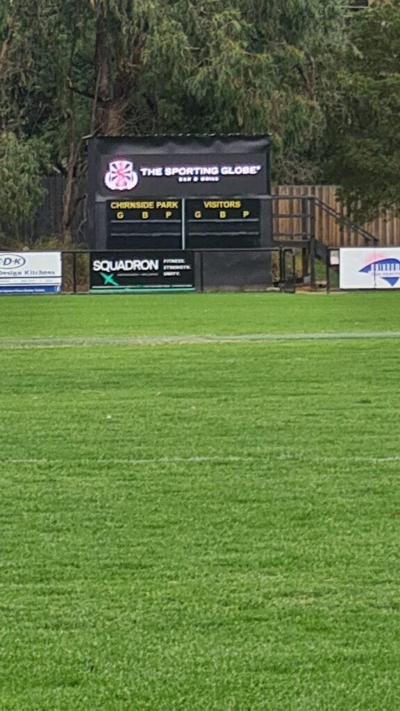 Outdoor LED scoreboard displaying live match information at a community sports ground